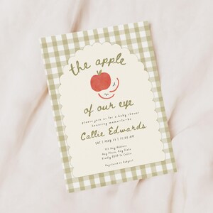 May include: A rectangular baby shower invitation card featuring a sage green and white gingham border. The center has a scalloped cream-colored panel with a red apple illustration and the text: the apple of our eye, please join us for a baby shower honoring mama-to-be Callie Edwards, SAT | MAY 23 | 11:00 AM, 123 Any Address, Any Place, Any State, Kindly RSVP to Callie, Registered on Babylist.