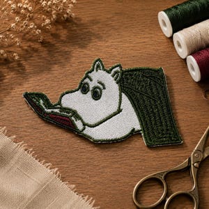 May include: An embroidered patch featuring a white, hippopotamus-like character with dark green outlines, reading an open book with a dark red cover. The character has large, dark eyes and is depicted in profile, with a dark green tail extending to the right.