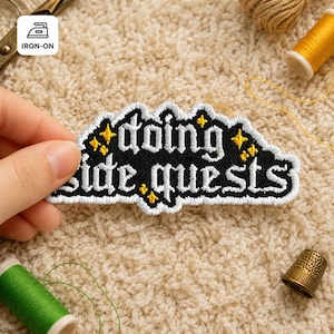 May include: Black iron-on patch with the words "doing side quests" in white gothic lettering. The patch has a white border and yellow star accents. The patch is held by a hand, with sewing supplies in the background.
