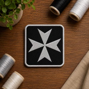 May include: A square embroidered patch featuring a white Maltese cross centered on a black background with a white stitched border. The patch has a clean, geometric design with sharp, pointed edges on the cross, creating a high-contrast visual.