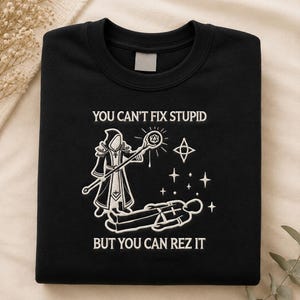 May include: Black sweatshirt with white graphic text and illustration. The text reads "YOU CAN'T FIX STUPID BUT YOU CAN REZ IT". The illustration depicts a robed figure with a staff and a person lying on the ground with stars.