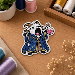 Op de afbeelding: Een geborduurde patch met een witte geitfiguur met hoorns, gekleed in een donkerblauwe tovenaarsgewaad met gouden sterren en een halve maan, die een klein glazen flesje met een roze drankje in een hoef vasthoudt.