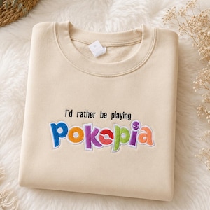 May include: Cream-colored sweatshirt with the embroidered text "I'd rather be playing Pokepia" in colorful letters. The sweatshirt has a crew neck and a tag on the collar.