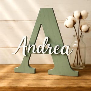 Può includere: Una lettera "A" in legno verde salvia con il nome "Andrea" in corsivo bianco. La lettera è su una superficie di legno, con un vaso di vetro con steli di cotone sullo sfondo. Un oggetto decorativo.