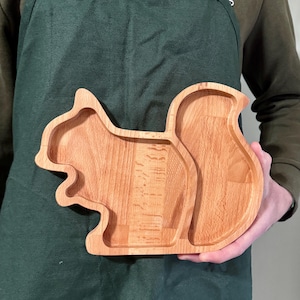Puede incluir: Una bandeja de servicio de madera de color marrón claro con forma de ardilla y una cola grande y curva. La bandeja presenta un acabado liso de veta de madera natural y un área interior rebajada para colocar objetos pequeños, aperitivos o decoraciones.