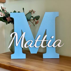 Può includere: Una grande lettera M in legno azzurro con il nome Mattia in corsivo bianco applicato sulla parte anteriore. La lettera presenta una finitura liscia e opaca e si regge in verticale, mostrando un design pulito con una tipografia chiara.