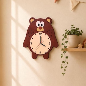 Puede incluir: Un reloj de pared con forma de oso marrón con una esfera beige claro. El reloj tiene manecillas negras y números negros en las posiciones de las 12, 3, 6 y 9 en punto. Los ojos y la nariz del oso son de color marrón claro. El reloj está montado en una pared beige claro.