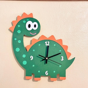 Op de afbeelding: Een groene wandklok in de vorm van een dinosaurus met oranje stekels langs de rug en staart. De wijzerplaat toont de witte cijfers 12, 3, 6 en 9, met zwarte wijzers. De dinosaurus heeft lichtgroene stippen en een glimlachend gezicht.