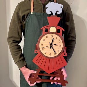 Puede incluir: Un reloj de madera rojo y marrón con forma de tren y una esfera de madera clara. El reloj tiene manecillas y números negros. El reloj está sostenido por un soporte de madera marrón. El reloj mide aproximadamente 30 cm de alto.