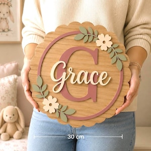 Può includere: Una targa circolare in legno con bordo smerlato, che misura 30 cm di diametro. La targa presenta una grande lettera G rosa scuro, il nome Grace in corsivo bianco e foglie verde chiaro e fiori color crema decorativi.