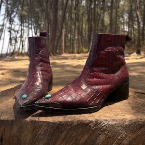 May include: A pair of dark burgundy ankle boots with a crocodile-embossed texture, featuring a pointed toe, a low block heel, and a small turquoise stone accent set into the metal-capped toe of each boot, with a side zipper closure.