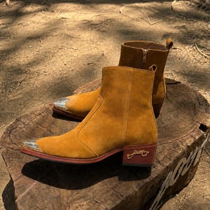 May include: A pair of tan suede ankle boots featuring pointed toes with silver-toned metal caps. The boots have a low block heel with a gold-toned cursive logo detail on the side and a side zipper closure with a gold-toned pull tab.
