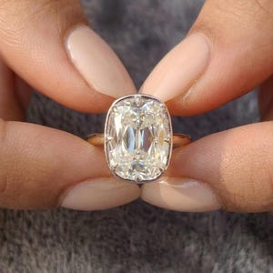 Peut inclure: Une bague avec une grande pierre précieuse transparente taille coussin, sertie dans une monture en métal poli. La pierre présente de multiples facettes qui reflètent la lumière, et l'anneau est un design métallique simple et lisse.