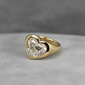 May include: A gold ring featuring a large, heart-shaped diamond. The diamond is set in a bezel setting, and the band is a smooth, polished gold. The ring is displayed on a soft, gray surface.
