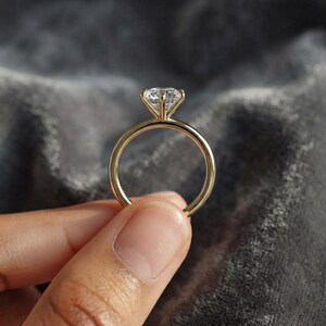 Könnte beinhalten: Ein goldener Ring mit einem einzelnen, rund geschliffenen, klaren Edelstein in einer Vier-Krappen-Fassung. Die Ringschiene ist glatt und poliert und zeigt ein klassisches Solitär-Design mit einer Fassung, die den Stein über dem Ring anhebt.