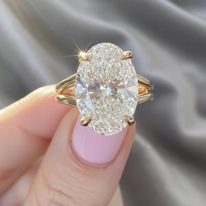 Op de afbeelding: Een gouden ring met een grote, ovaal geslepen diamant. De heldere diamant schittert, vastgehouden door vier klauwen. De ring heeft een split-schacht ontwerp. De ring wordt tegen een grijze achtergrond gehouden.