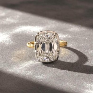 Puede incluir: Un anillo de diamantes con una piedra de talla rectangular y una banda de oro. El diamante está engastado en un halo de platino u oro blanco. El anillo se muestra sobre una superficie gris brillante.