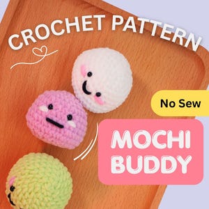 Może przedstawiać: Trzy szydełkowe mochi w kolorze białym, różowym i zielonym, każdy z uśmiechniętą twarzą. Obraz zawiera tekst "CROCHET PATTERN" i "MOCHI BUDDY" na drewnianej tacy.