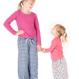 May include: Two girls, one wearing a pink long-sleeved shirt and blue and white checkered pants, and the other wearing a pink long-sleeved shirt and a colorful floral skirt, are holding hands and looking at each other.