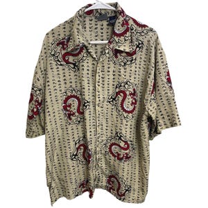 Pode incluir: Camisa bege de manga curta com um design de dragão vermelho. A camisa apresenta um padrão repetido de dragões vermelhos com detalhes em preto e caracteres japoneses. A camisa está pendurada em um cabide branco.