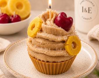Könnte beinhalten: Eine cupcake-förmige Kerze mit brennendem Docht, mit beigefarbenem Zuckerguss, der mit klaren zuckerartigen Kristallen, zwei gelben Ananasring-Dekorationen und einer roten Kirsche verziert ist. Die Kerze steht in einer braunen geriffelten Basis.