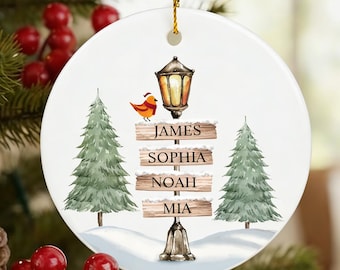 May include: A round, white Christmas ornament with a gold string, hanging on a Christmas tree. The ornament features a winter scene with a lamp post, bird, trees, and names on signs: James, Sophia, Noah, and Mia.
