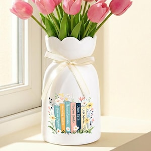 May include: A white vase with a scalloped rim holds a bouquet of pink tulips. The vase is decorated with a floral design and a ribbon. The vase is sitting on a windowsill next to a window.