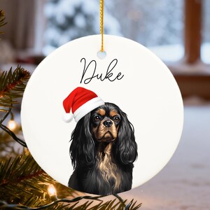 Puede incluir: Adorno navideño redondo y blanco con un retrato de un Cavalier King Charles Spaniel con un gorro de Papá Noel rojo. El nombre "Duke" está escrito encima. El perro tiene pelo negro y marrón. Una cuerda dorada está adjunta.