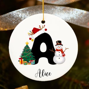 Può includere: Ornamento natalizio rotondo e bianco con la lettera nera "A", un albero di Natale, un pupazzo di neve e un cappello di Babbo Natale. Il nome "Alice" è scritto sotto. Appeso a un filo d'oro.