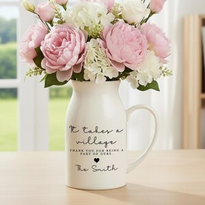 Könnte beinhalten: Eine weiße Keramikvase mit Henkel, gefüllt mit rosa und weißen Blumen. Die Vase trägt den Text "It takes a village, thank you for being a part of ours, The Smith" in schwarzer Schrift. Die Vase steht auf einem Holztisch.
