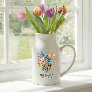 May include: A white ceramic pitcher with a handle, filled with colorful tulips. The pitcher is decorated with a floral bouquet and the text "THE GALLING EST. 2026". The flowers include pink, purple, yellow, and white tulips.