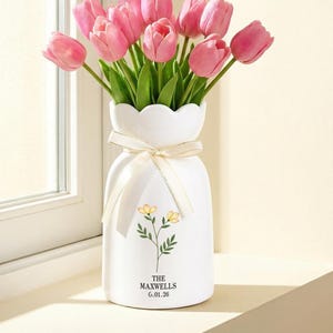 May include: A white ceramic vase with a scalloped rim displays a bouquet of pink tulips. The vase is decorated with a floral design and a cream ribbon. The text "THE MAXWELLS 6.01.26" is printed on the vase.