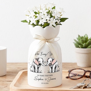 May include: A white ceramic vase with a floral arrangement, featuring two elephants with intertwined trunks. The vase has text that reads "The Ivory Years" and "14 Years Together". A white mug, glasses, and keys are also visible.