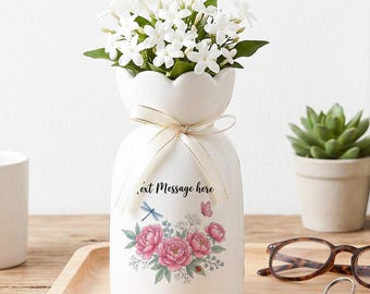 May include: A white ceramic vase with a scalloped rim, tied with a thin cream ribbon. The vase features a floral print of pink peonies, a blue dragonfly, and a small butterfly, with the black printed text "Insert Message here" above the flowers.