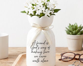Puede incluir: Un jarrón de cerámica blanca con borde festoneado, atado con una cinta fina de color crema, que contiene flores de jazmín blancas. El jarrón presenta el texto impreso en negro: A friend is God's way of making sure we never walk alone. Acabado liso.