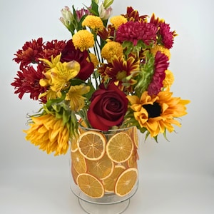 May include: A vibrant floral arrangement in a clear glass vase decorated with dried orange slices. The bouquet features sunflowers, roses, and chrysanthemums in shades of yellow, red, and burgundy. The vase sits on a clear, pedestal base.