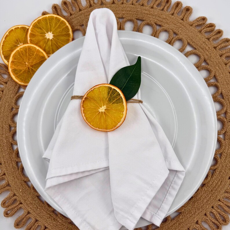 May include: A white folded napkin on a white plate, adorned with a dried orange slice and a green leaf, tied with twine. The plate rests on a brown woven placemat, with additional orange slices nearby. A rustic, natural table setting.