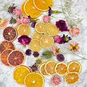 May include: Overhead shot of dried citrus fruit slices, including oranges, blood oranges, and lemons, arranged with dried pink and purple roses, and sprigs of white flowers on a marble surface.