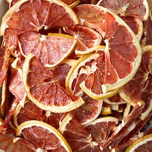 May include: Close-up of dried grapefruit slices. The slices are a deep red color with a yellow rind. The texture of the fruit is visible, with the segments and seeds clearly defined. The slices are arranged in a pile, creating a visually appealing display.