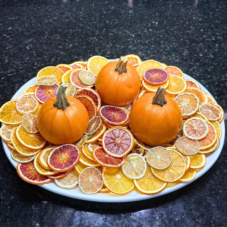 May include: A white oval platter holds three small orange pumpkins surrounded by dried orange and citrus slices. The arrangement features a variety of colors, including orange, yellow, and red, creating a festive and autumnal display.