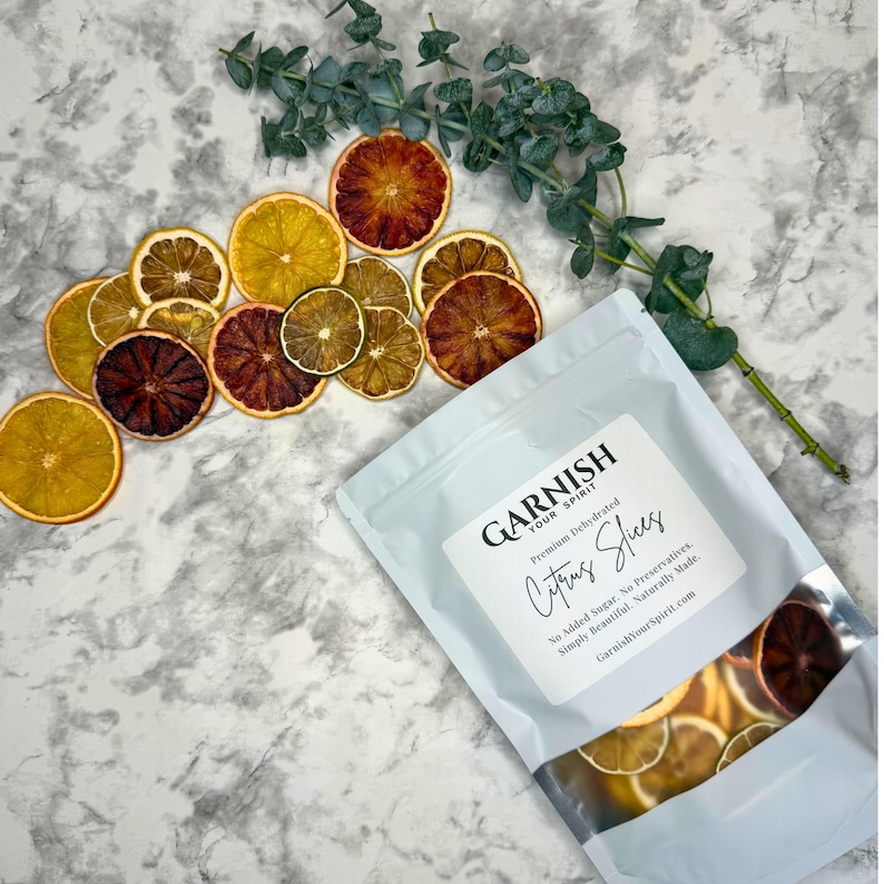 May include: A white bag of dehydrated citrus slices with the text "GARNISH YOUR SPIRIT" and "Citrus Slices". The bag is next to a collection of dried orange, lemon, and lime slices, and a sprig of eucalyptus.