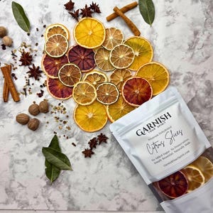 May include: Dehydrated citrus slices in a white bag with the text "GARNISH YOUR SPIRIT Citrus Slices". The image also includes star anise, cinnamon sticks, nutmeg, and bay leaves. The slices are orange, yellow, and red.