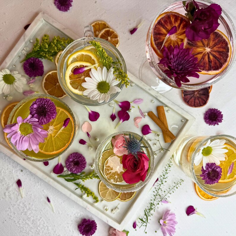 May include: Overhead shot of several clear glasses and a tray with floral garnishes. The drinks contain citrus slices, flowers, and herbs. Dried orange slices and cinnamon sticks are also present, with scattered flower petals.