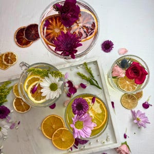 May include: Overhead shot of several cocktails garnished with flowers and citrus slices. The drinks are in clear glasses, some with sugared rims. Dried orange slices and flower petals are scattered around the glasses on a white surface.