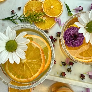 May include: Two cocktails with orange slices and flowers, garnished with sugar rims. One glass has a white daisy, the other a purple chrysanthemum. Dried orange slices and floral accents are scattered around the glasses on a white marble surface.