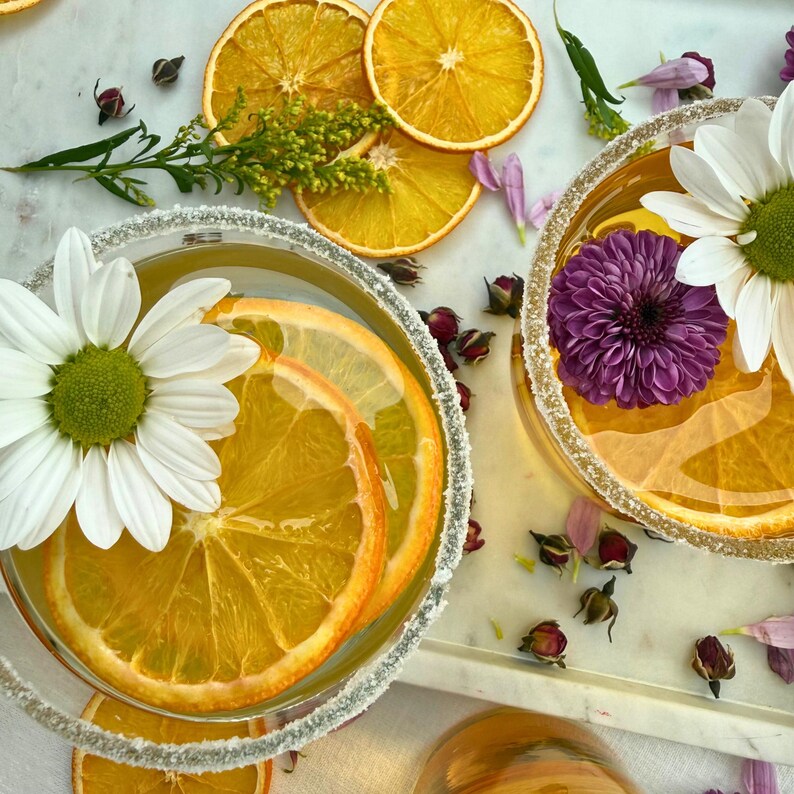 May include: Two cocktails with orange slices and flowers, garnished with sugar rims. One glass has a white daisy, the other a purple chrysanthemum. Dried orange slices and floral accents are scattered around the glasses on a white marble surface.