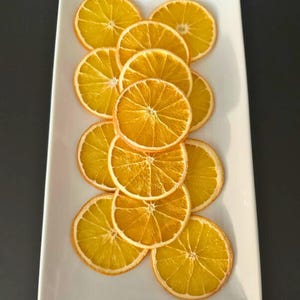 May include: A rectangular white platter displays a collection of dried orange slices. The slices are arranged in a vertical pattern, showcasing their circular shape and vibrant orange color. The dried oranges have a textured appearance.