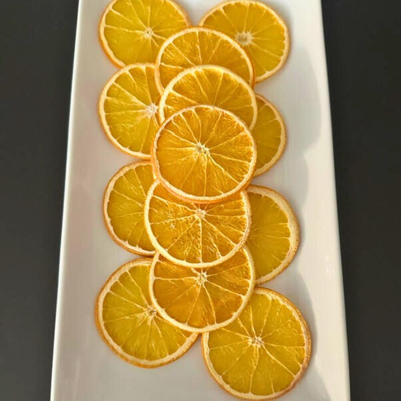 May include: A rectangular white platter displays a collection of dried orange slices. The slices are arranged in a vertical pattern, showcasing their circular shape and vibrant orange color. The dried oranges have a textured appearance.