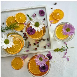 May include: A top-down view of four clear glasses filled with a golden liquid, orange slices, and flowers. The glasses are garnished with sugar rims and sit on a white surface. A white tray with orange slices and flowers is also present.