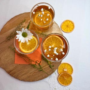 May include: Three clear glasses filled with a golden liquid, garnished with orange slices and small white flowers. The rims of the glasses are sugared. The drinks sit on a wooden board with an orange napkin and dried orange slices.